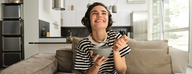 Close-up von glücklicher lachender Frau, die lächelnd vor dem Fernseher isst und eine Schüssel und einen Löffel trägt
