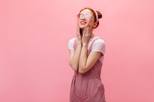 Closeup Portrait der jungen Dame mit Stirnband und in rosa Brille Frau im Overall posiert mit einem Lächeln auf rosa Hintergrund