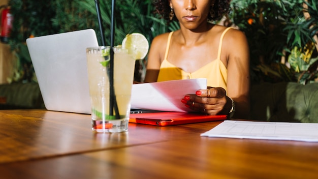 Cocktailglas auf Tabelle vor der jungen Frau, die das Dokument im Restaurant überprüft