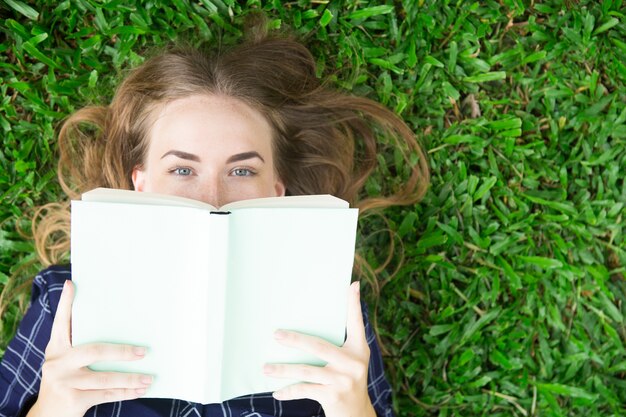 Content Girl liegt auf Gras und versteckt sich hinter Buch