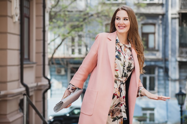 Coole attraktive stilvolle lächelnde Frau, die Stadtstraße in rosa Mantelfrühlingsmodetrend hält Geldbörse geht