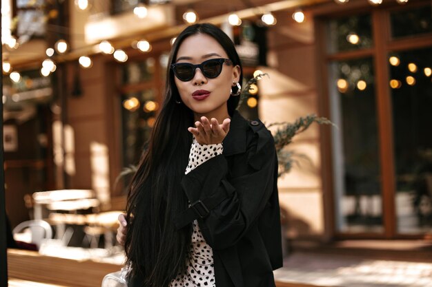 Coole brünette Frau mit Sonnenbrille bläst Kuss Portrait der asiatischen attraktiven Dame im schwarzen Trenchcoat Hübsches Mädchen posiert draußen in guter Laune