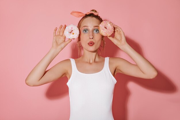 Coole Dame mit hellem Make-up und modernen Ohrringen im weißen trendigen Badeanzug posiert mit zwei Donuts auf isoliertem rosa Hintergrund