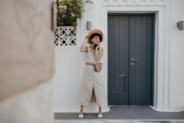 Coole junge brünette, lockige Frau in weißem Outfit und Hut lehnt sich draußen an die Wand Charmante Dame im sommerlichen Midikleid und Boater posiert in der Nähe einer grauen Holztür