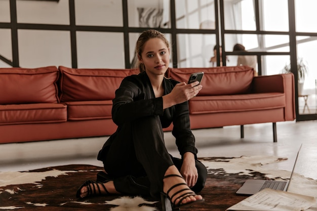 Coole junge Frau im stylischen schwarzen Seidenanzug sitzt auf dem Teppich im Wohnzimmer und hält ein Telefon