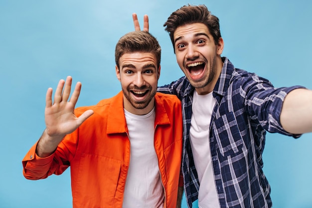 Coole jungs in stylischen outfits haben spaß und lächeln breit auf blauem hintergrund guy im karierten hemd macht seinem freund hasenohren