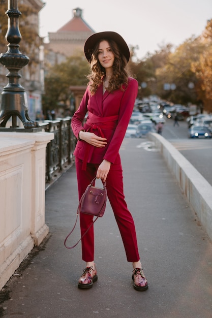 Coole schöne stilvolle Frau im lila Anzug, die in Stadtstraße, Frühlingssommer-Herbstsaison-Modetrend trägt Hut trägt und Geldbörse hält