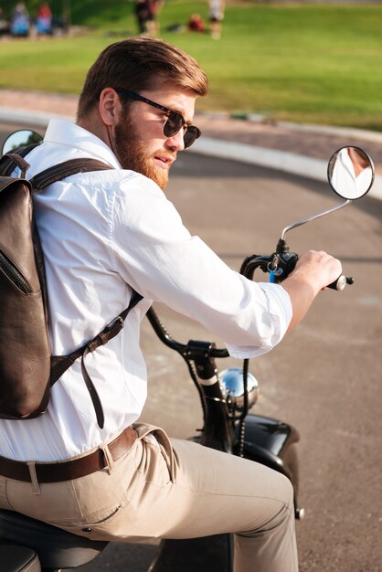 Cooler bärtiger Mann in Sonnenbrille mit Rucksack fährt auf modernem Motorrad im Freien und schaut weg