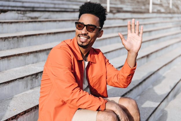 Cooler, fröhlicher dunkelhäutiger Mann mit Sonnenbrille und orangefarbener Jacke lächelt, winkt zur Begrüßung und sitzt auf der Treppe