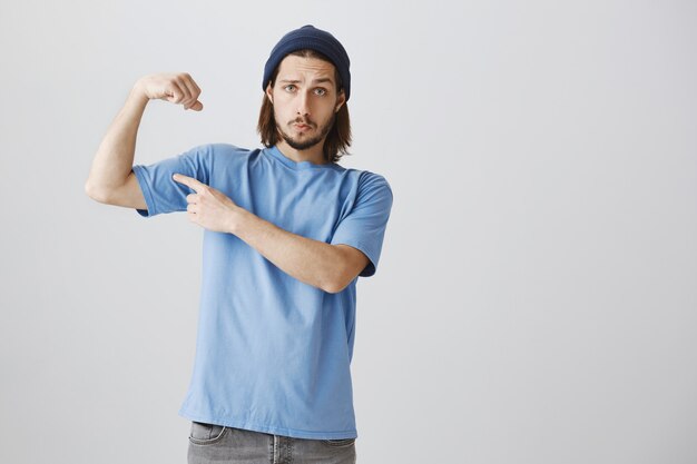 Cooler Hipster-Typ in blauem T-Shirt und Beanie-Flex-Bizeps, der mit starken Muskeln prahlt