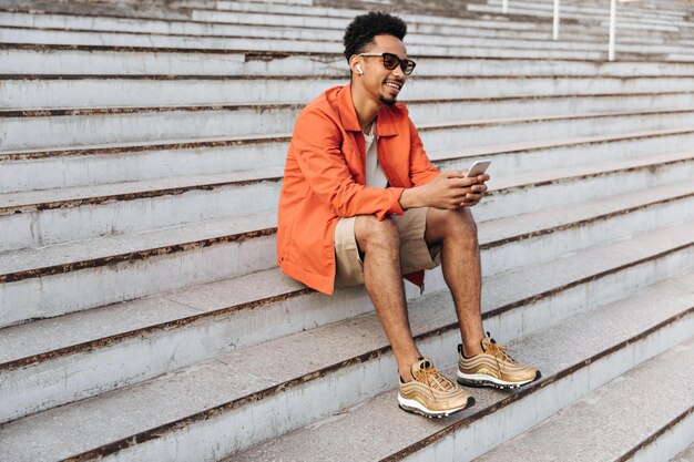 Cooler junger lockiger dunkelhäutiger Mann in orangefarbener Jacke und beigen Shorts sitzt draußen auf der Treppe Fröhlicher Kerl mit Sonnenbrille hält Telefon