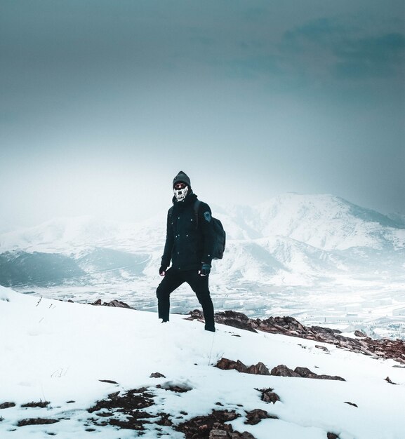 Cooler Wanderer in einer Maske und dunkler Kleidung, die allein auf einem schneebedeckten steilen Hügel in den Bergen stehen