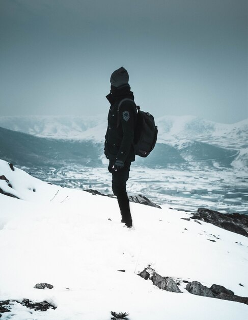 Cooler Wanderer in einer Maske und dunkler Kleidung, die allein auf einem schneebedeckten steilen Hügel in den Bergen stehen