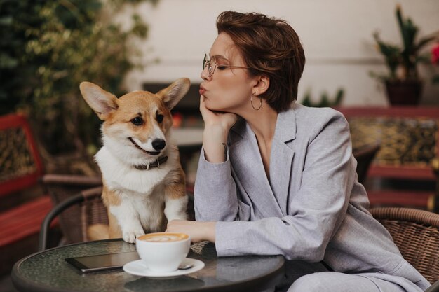 Cooles Mädchen im grauen Anzug küsst ihren Hund und friert im Straßencafé Hübsche dunkelhaarige Frau in Jacke, die draußen mit Corgi ruht