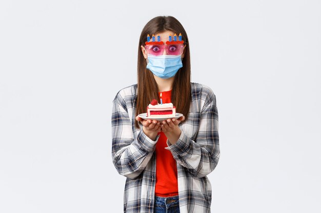 Coronavirus-Ausbruch, Lebensstil während der sozialen Distanzierung und Feiertagsfeierkonzept. Überraschtes oder überfallenes süßes Mädchen mit Brille und medizinischer Maske, das Geburtstagskuchen hält, verwirrt, wie Kerze bläst?