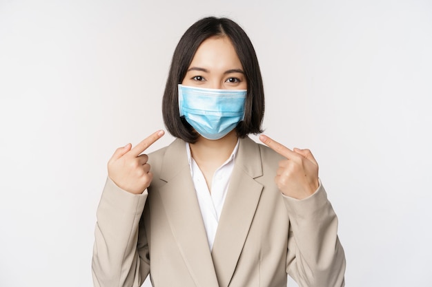 Coronavirus und Geschäftsleute Konzept Asiatische Unternehmerin, die mit dem Finger auf eine medizinische Gesichtsmaske am Arbeitsplatz zeigt, die über weißem Hintergrund steht
