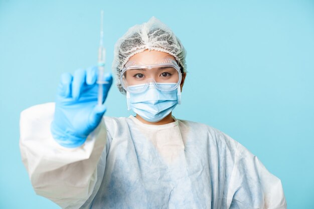 Coronavirus und Gesundheitskonzept. Asiatische Ärztin oder Krankenschwester, zeigt Spritze mit Impfstoff gegen Covid-19, Grippe, stehend in persönlicher Schutzausrüstung, blauer Hintergrund