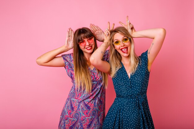 Coupé des besten Freundes, der Spaß hat, Hasenohren durch ihre Hände imitiert, bunte Sommerkleider und Sonnenbrille, rosa Wand, Partyzeit trägt.