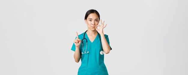 Covid-19, Beschäftigte im Gesundheitswesen, Pandemiekonzept. Ernst aussehende, besorgte asiatische Krankenschwester, Arzt bittet um Geheimhaltung, schüttelt den Finger und zeigt Mundsiegel, Geste mit zuckenden Lippen