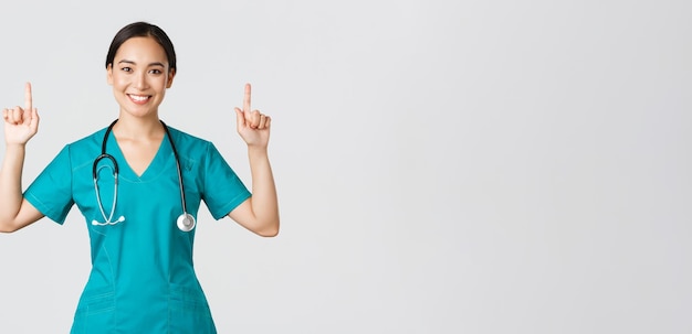 Covid-19, Beschäftigte im Gesundheitswesen, Pandemiekonzept. Glücklich lächelnde asiatische Ärztin, Krankenschwester in Scrubs, die Werbung zeigt, Ankündigung macht. Arzt zeigt mit den Fingern auf das Banner.