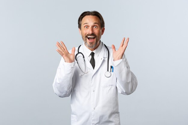 Covid-19, Coronavirus-Ausbruch, Gesundheitspersonal und Pandemiekonzept. Aufgeregt glücklicher männlicher Arzt, der großartige Neuigkeiten erzählt, die Hände hebt und begeistert lächelt und auf eine tolle Promo reagiert