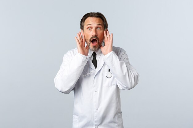 Covid-19, Coronavirus-Ausbruch, Gesundheitspersonal und Pandemiekonzept. Schockiert aufgeregter männlicher Arzt im weißen Kittel, der jemanden anruft und laut mit den Händen in der Nähe des geöffneten Mundes schreit