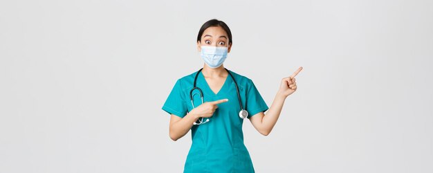 Covid-19, Coronavirus-Krankheit, Konzept der Beschäftigten im Gesundheitswesen. Begeisterte glückliche asiatische Ärztin, Ärztin oder Krankenschwester in medizinischer Maske und Peelings, die auf die obere rechte Ecke zeigt, zeigt tolle Promo