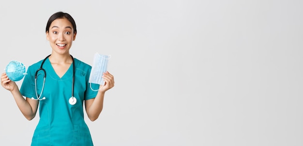 Covid-19, Coronavirus-Krankheit, Konzept der Mitarbeiter im Gesundheitswesen. Amüsierte, lächelnde asiatische Krankenschwester, die in Peelings mit Atemschutzmaske und medizinischer Maske interagiert, erhält persönliche Schutzausrüstung.