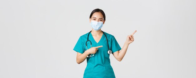 Covid-19, Coronavirus-Krankheit, Konzept der Mitarbeiter im Gesundheitswesen. Angenehme asiatische Ärztin, medizinische Angestellte in Scrubs und Maske, die mit dem Finger in die obere rechte Ecke zeigt und Werbung zeigt.