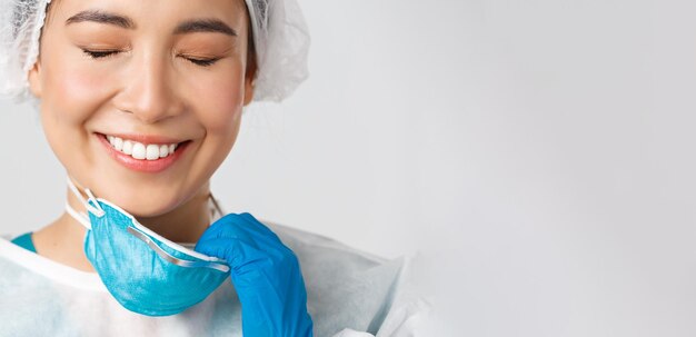 Covid-19, Coronavirus-Krankheit, Konzept der Mitarbeiter im Gesundheitswesen. Lächelnde erleichterte asiatische Ärztin nimmt Atemschutzmaske ab und hat Hautschäden durch persönliche Schutzausrüstung, weißer Hintergrund