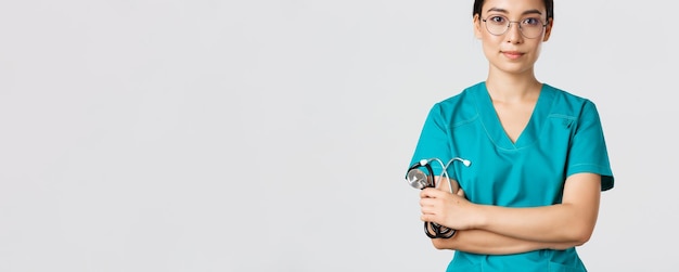 Covid-19, Coronavirus-Krankheit, Konzept für Gesundheitspersonal. Zuversichtlich lächelnder professioneller asiatischer Arzt mit Brille, Brust mit verschränkten Armen, trägt Peelings und hält Stethoskop, weißer Hintergrund.