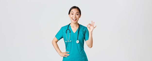 Covid-19, Gesundheitspersonal, Pandemiekonzept. Überraschte und glückliche asiatische Ärztin, Krankenschwester in Peelings, die eine gute Geste zeigt und erstaunt lächelt, gute Arbeit loben, jemandem zustimmen.