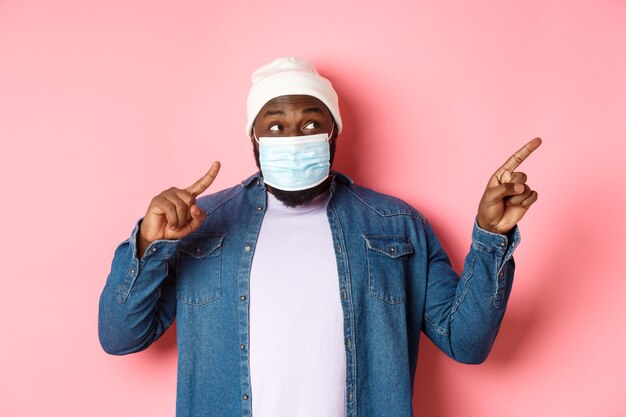 Covid-19, Lifestyle- und Lockdown-Konzept. Amüsierter schwarzer Mann in medizinischer Maske, der direkt auf den Kopierraum schaut und zeigt, auf rosafarbenem Hintergrund steht.