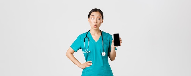 Covid-19, medizinisches Personal und Online-Medizinkonzept. Aufgeregte und erstaunte asiatische Krankenschwester, Ärztin sieht überrascht aus, während sie Handybildschirm, Internetberatungs-App, weißen Hintergrund zeigt.