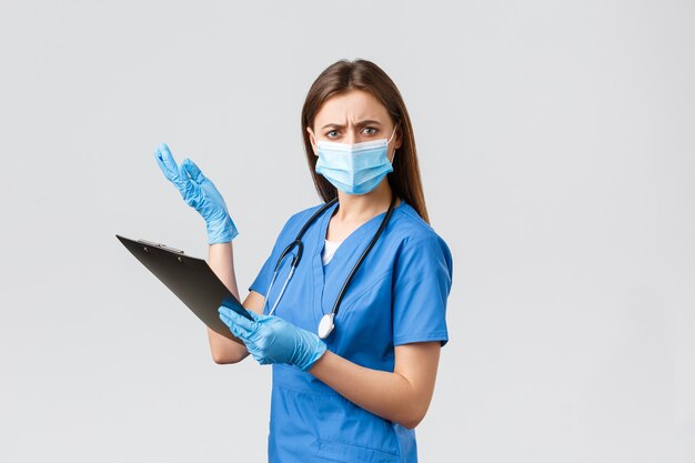 Covid-19, Prävention von Viren, Gesundheit, Gesundheitspersonal und Quarantänekonzept. Unzufriedene und belästigte Krankenschwester oder Ärztin in blauen Kitteln und medizinischer Maske, die Unsinn in der Zwischenablage liest