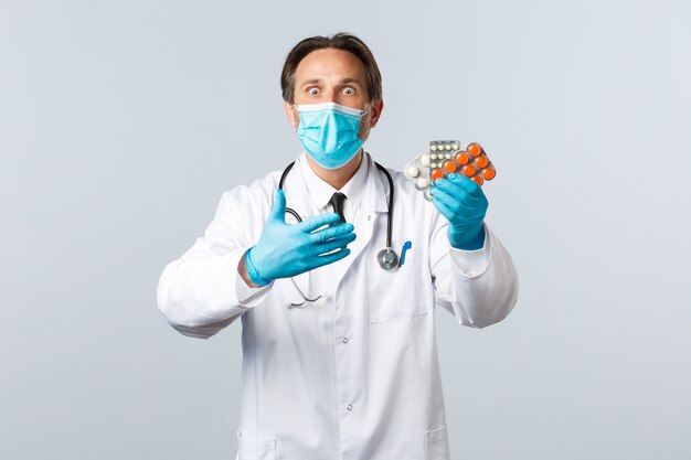 Covid-19, Prävention von Viren, Gesundheitspersonal und Impfkonzept. Aufgeregter und schockierter Arzt in medizinischer Maske und Handschuhen reagiert auf neue Medikamente und zeigt Pillen, weißen Hintergrund