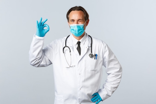 Covid-19, Prävention von Viren, Gesundheitspersonal und Impfkonzept. Fröhlicher aufgeregter Arzt in medizinischer Maske und Handschuhen, zwinkert und zeigt ein OK-Zeichen, garantiert Qualität, weißer Hintergrund.