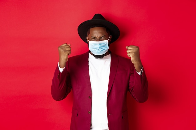 Covid-19, quarantäne- und feiertagskonzept. fröhlicher afroamerikanischer mann, der geballte faust zeigt und sich über den sieg freut, das ziel erreicht und eine medizinische maske aus coronavirus trägt