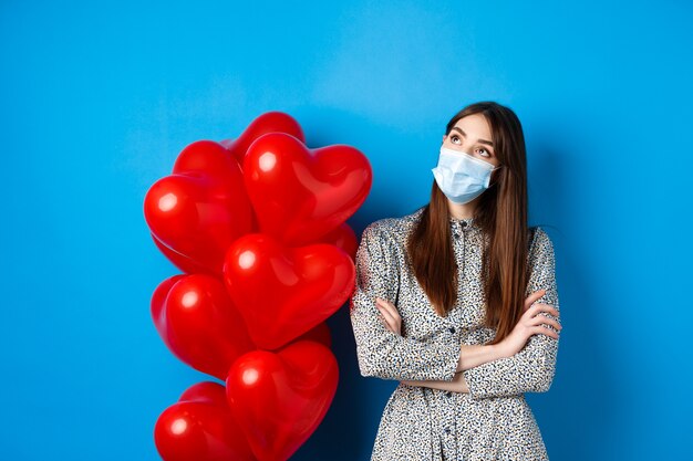 Covid-19, Quarantäne und Gesundheitskonzept. Verträumtes schönes Mädchen in Gesichtsmaske und Kleid, Blick auf die obere linke Ecke nachdenklich, in der Nähe von Valentinstagballons stehen, blauer Hintergrund