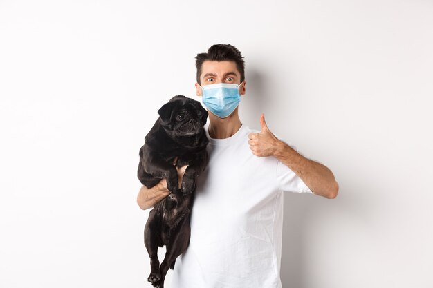 Covid-19, Tiere und Quarantänekonzept. Junger Mann in medizinischer Maske, der einen süßen schwarzen Mops-Hund hält, Daumen nach oben zeigt, wie und genehmigt, auf weißem Hintergrund stehend