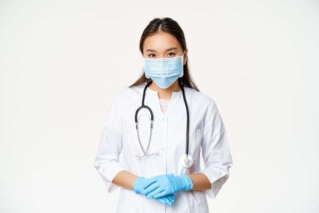 Covid-19 und Gesundheitskonzept. Junge asiatische Ärztin in medizinischer Maske, Gummihandschuhen und Klinikuniform, die bereit ist, zu helfen, zuhörender Patient, weißer Hintergrund.