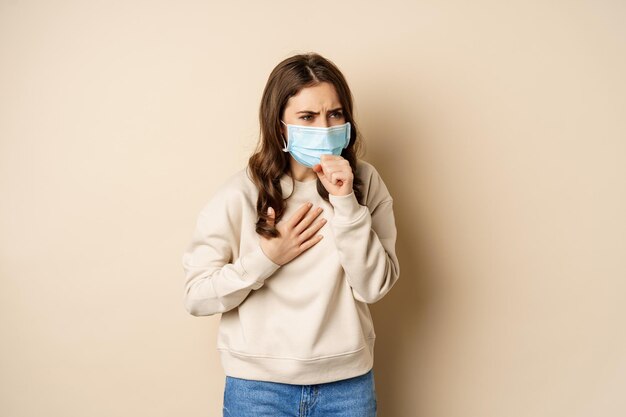 Covid-19 und Gesundheitskonzept. Kranke Frau in medizinischer Gesichtsmaske hustet, fühlt sich krank mit saurem Hals und steht auf beigem Hintergrund