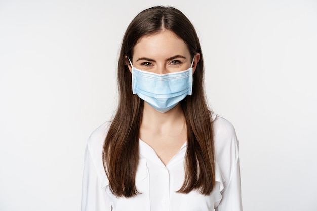 Covid-19 und Pandemiekonzept. Junge Bürofrau, die während der sozialen Distanzierung des Coronavirus eine medizinische Maske trägt und über weißem Hintergrund steht
