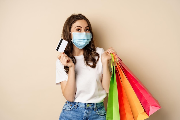 Covid-Pandemie und Lifestyle-Konzept Junge Frau posiert in medizinischer Gesichtsmaske mit Einkaufstüten aus...