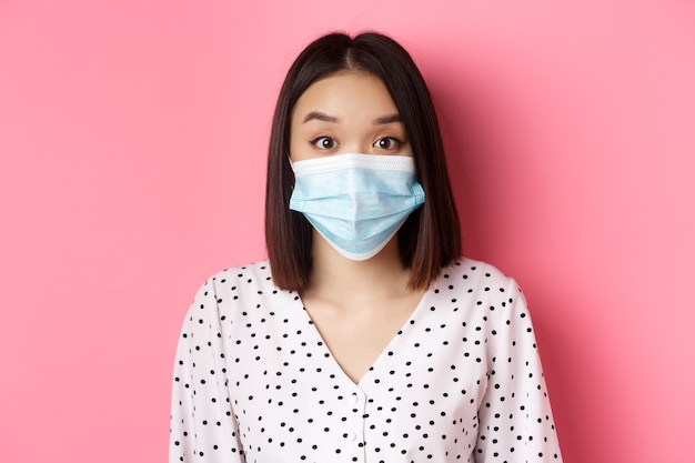 Covid-Pandemie- und Lifestyle-Konzept überraschte asiatische Frau in Gesichtsmaske, die Augenbrauen hochzieht und auf ...