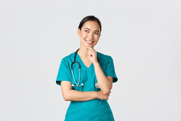 Covid19 Gesundheitspersonal Pandemiekonzept Lächelnd erfreut attraktive asiatische Ärztin in Scrubs, die in die obere linke Ecke schaut und denkt, dass sie eine Idee hat, nachdenklich weißen Hintergrund zu stehen