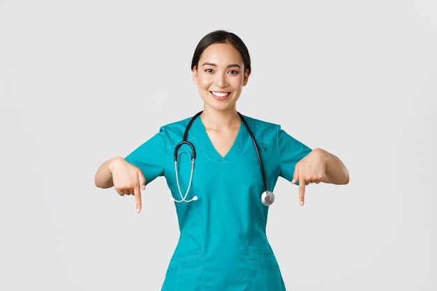 Covid19 Gesundheitspersonal Pandemiekonzept Lächelnde, angenehme asiatische Ärztin, Therapeutin oder Ärztin in Scrubs mit Stethoskop, die mit dem Finger nach unten zeigen, zeigen Klinikbanner