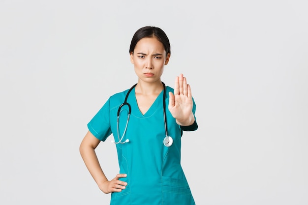 Covid19 Gesundheitspersonal Pandemiekonzept Wütend ernsthaft aussehende asiatische Ärztin oder Krankenschwester in Peelings, die unzufrieden die Stirn runzeln, strecken die Hand aus, um zu zeigen, dass sie nicht einverstanden sind, zu verbieten oder zu verbieten