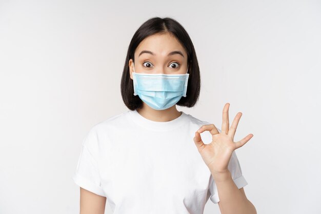 Covid19 Gesundheitswesen und medizinisches Konzept Beeindruckte asiatische Frau in medizinischer Maske, die erstaunt aussieht und ein ok-ok-Zeichen zeigt, das auf weißem Hintergrund steht