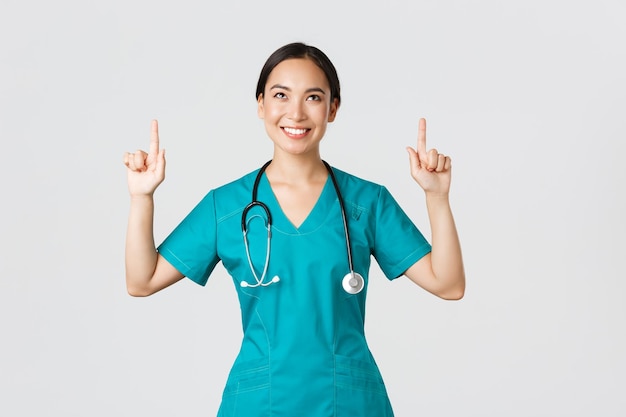 Covid19 Healthcare Workers Pandemiekonzept Lächelnde, schöne asiatische Krankenschwesterärztin in Scrubs, die mit einem zufriedenen Grinsen nach oben zeigt und gerne ein fantastisches Promo-Angebot auf weißem Hintergrund zeigt